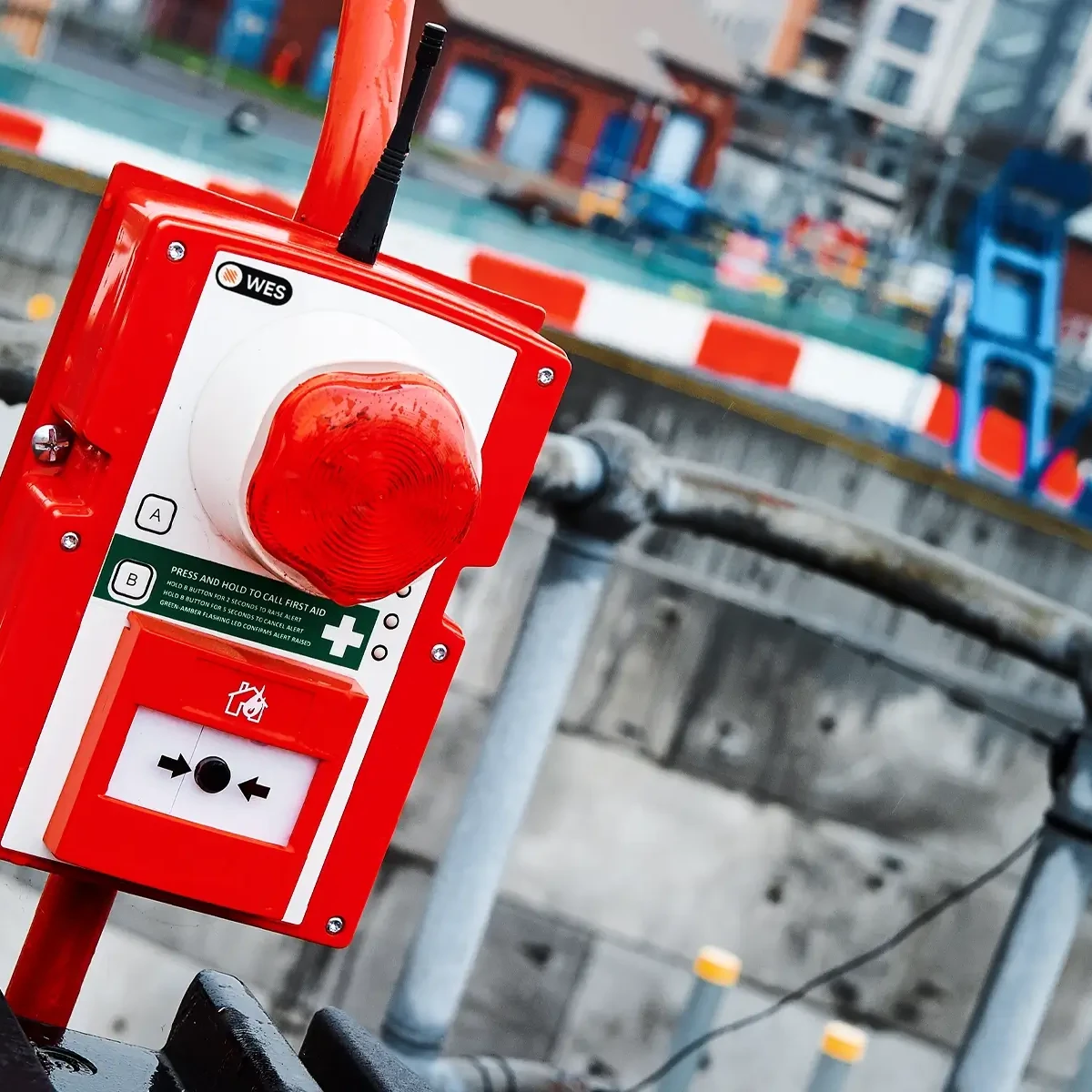 A WES3 Strobe and Sounder Call Point, a red and white wireless fire alarm and medical alert device with a large red button, installed outdoors on a temporary structure at a construction site.